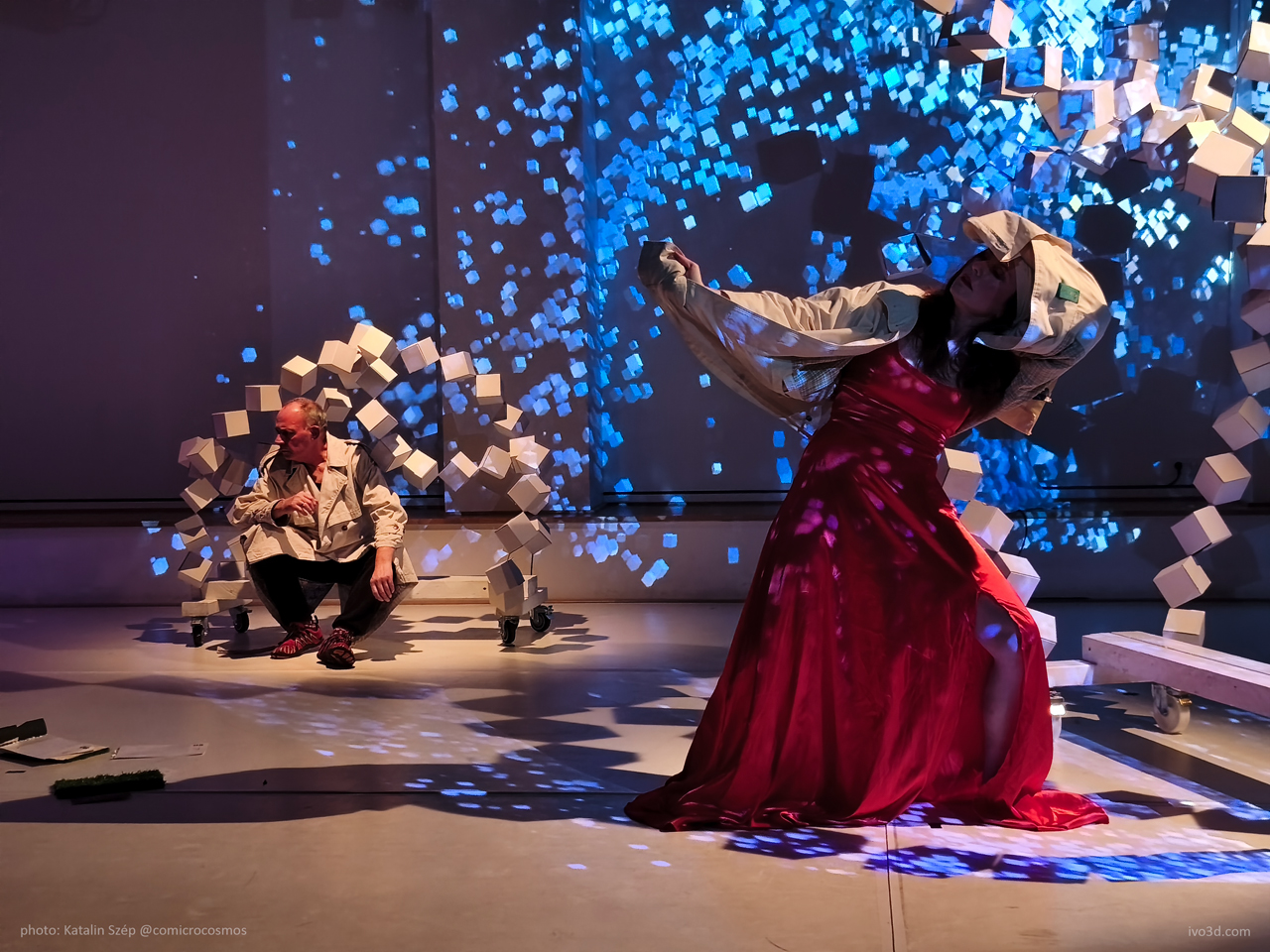 A male performer in a beige coat sits on the floor amidst a creative backdrop of floating blue digital cubes. A female performer in a red dress dances dynamically, showcasing expressive movement. The stage is illuminated with colorful lighting (projection), enhancing the artistic ambiance.