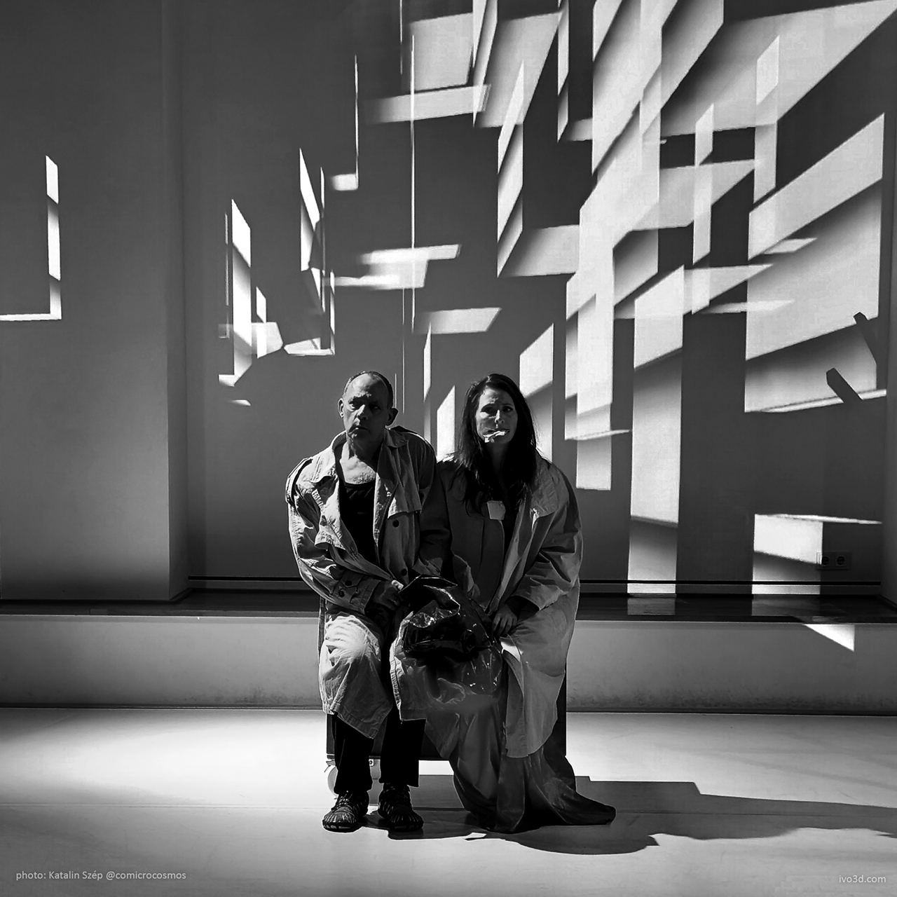 A black and white image of two seated performers in light trench coats, positioned in front of a wall casting geometric shadows of the video-projection