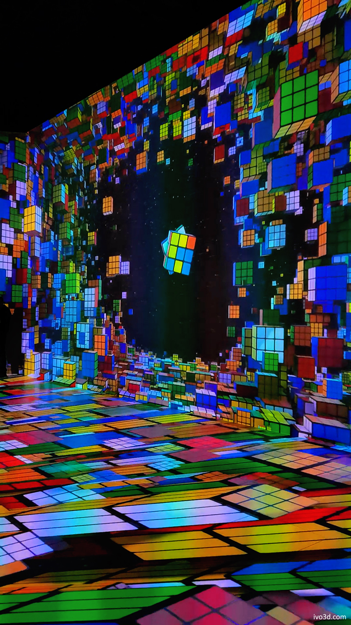 A vibrant, immersive room filled with colorful projections of Rubik's cubes on the walls and floor, creating a dynamic and playful atmosphere.